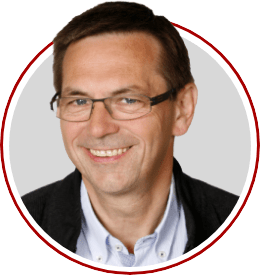 Ein Mann mit dunklen Haaren, einer Brille und schickem Hemd und Jacket lächelt freundlich